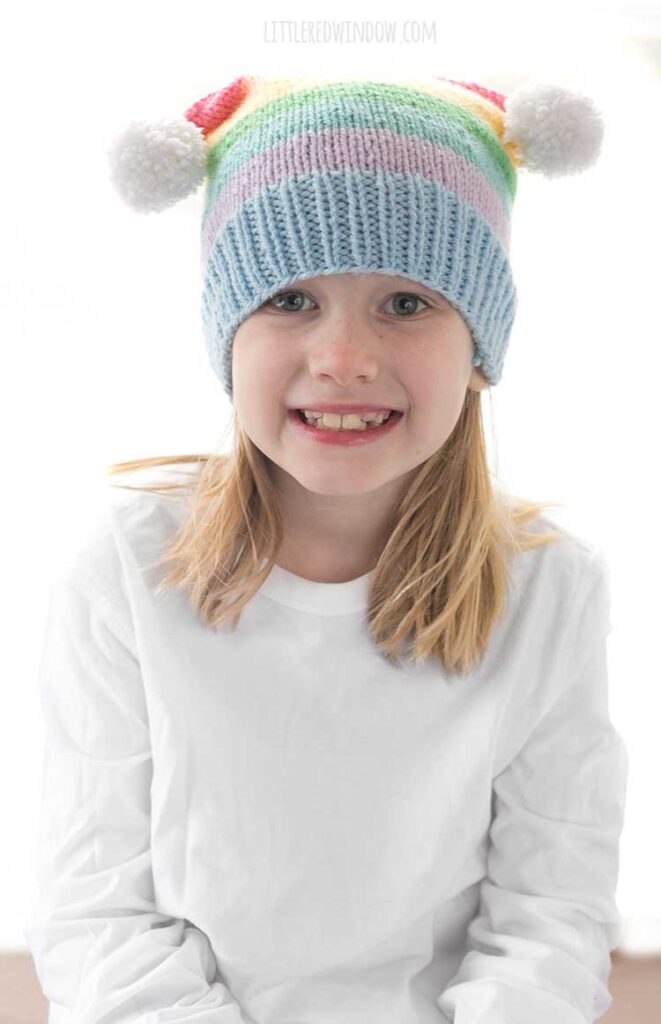 girl in white shirt wearing a rainbow striped hat with two white pom poms on the top corners looking at the camera and smiling