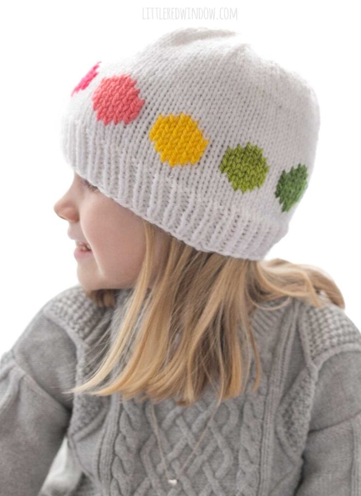 child in gray cable knit sweater wearing a white knit hat with warm rainbow colored large polka dots in a line around the middle looking off to the left