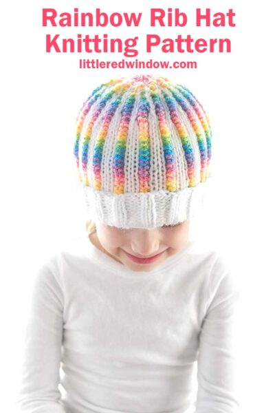 A child wearing a white sweater is smiling while looking down, showcasing the rainbow rib hat with a colorful pattern. The text above reads, "Rainbow Rib Hat Knitting Pattern littleredwindow.com.