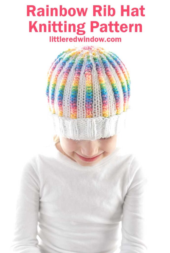 A child wearing a white sweater is smiling while looking down, showcasing the rainbow rib hat with a colorful pattern. The text above reads,
