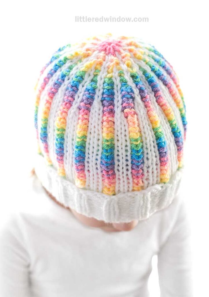 A child wearing a vibrant rainbow rib hat looks down, dressed in a white long-sleeve shirt against a plain white background.