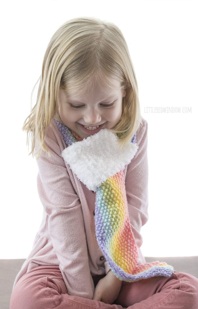 little girl looking down and wearing a rainbow striped knit scarf in front of a white background