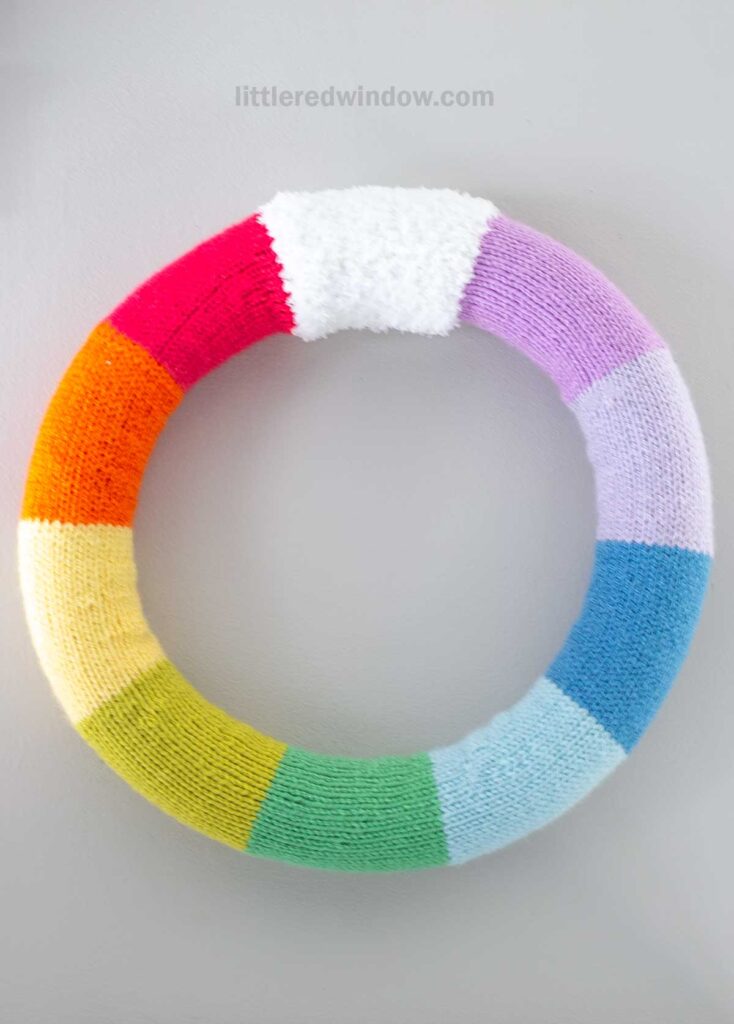 Round wreath covered in knitted fabric featuring a rainbow pattern with sections of red, orange, yellow, green, blue, purple, and white against a plain background.