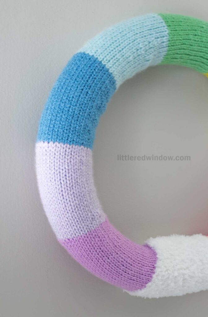Knitted wreath with colorful sections in blue, green, white, pink, and purple, displayed against a light gray background.