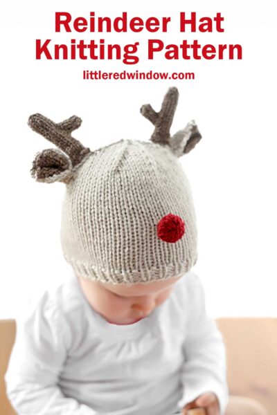 A baby in a reindeer hat with antlers and a red pom-pom nose is looking down, dressed in white. The text above reads, "Reindeer Hat Knitting Pattern, littleredwindow.com.
