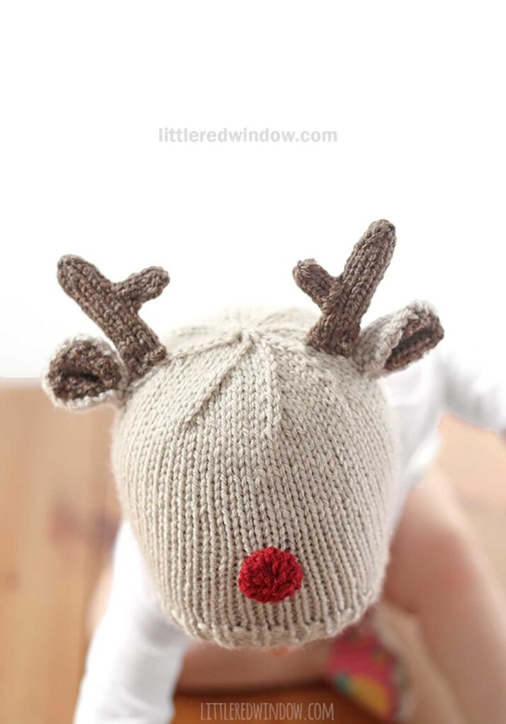 A baby in a charming reindeer hat, complete with antlers and a red nose, crawls on a wooden surface against a white background. The text