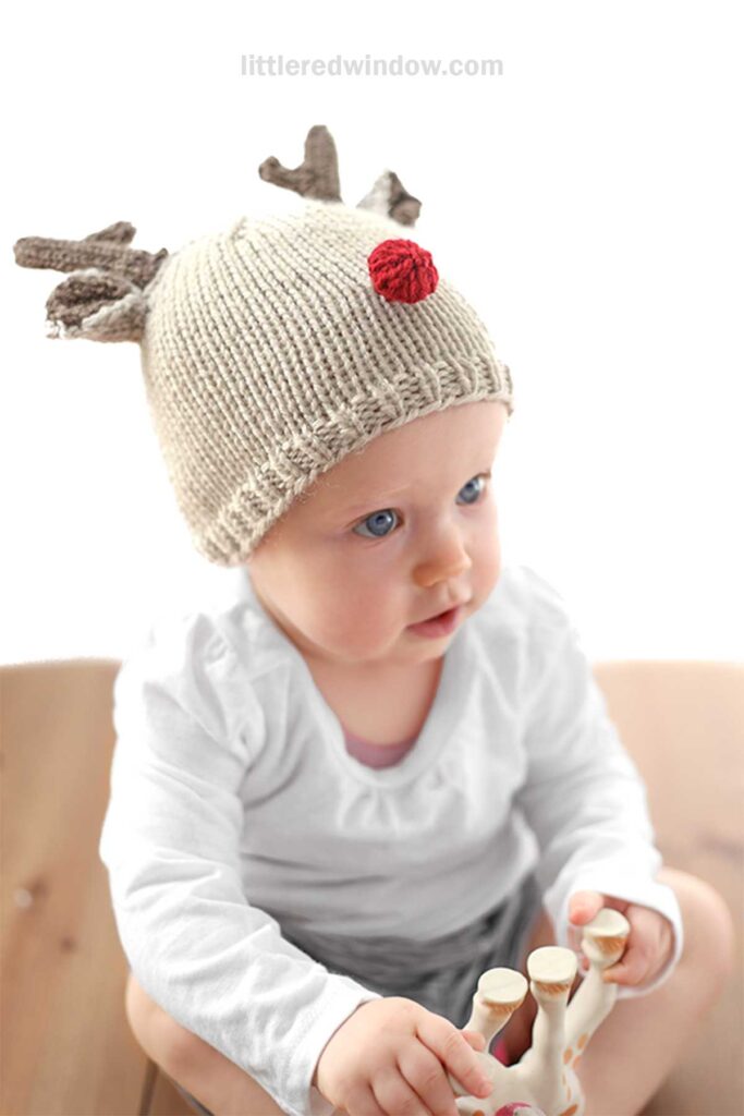 A baby with a charming reindeer hat adorned with antlers and a red nose sits on a wooden surface. Dressed in a white long-sleeve shirt, the little one holds a toy giraffe, adding to the adorable scene.