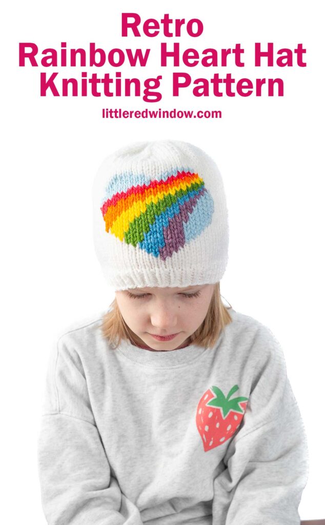 Child wearing a white knit hat with a rainbow heart design. The child is also wearing a gray sweatshirt with a strawberry patch. Text above reads