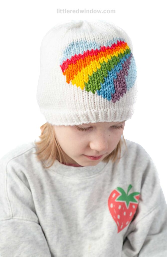 A child in a cozy gray sweater with a strawberry graphic looks down thoughtfully, their head adorned with a white knit hat showcasing an inspiring rainbow heart knitting pattern. The simple white background adds to the charm of this delightful ensemble.