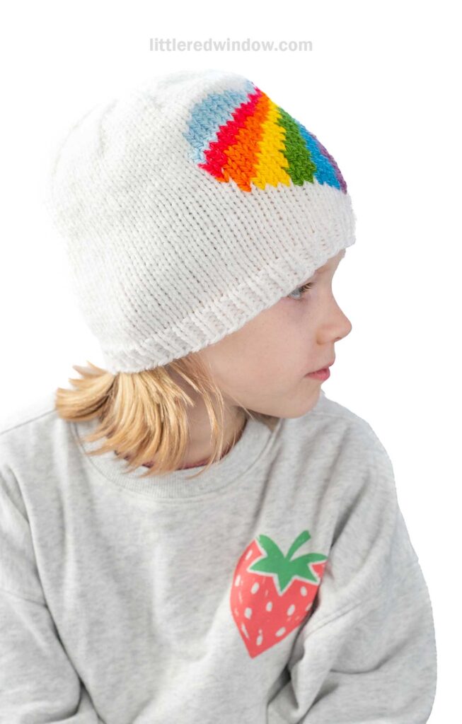 A child wearing a white knit hat with a rainbow heart pattern is facing to the side. The child is also wearing a gray sweatshirt with a strawberry design on it. The background is plain white.