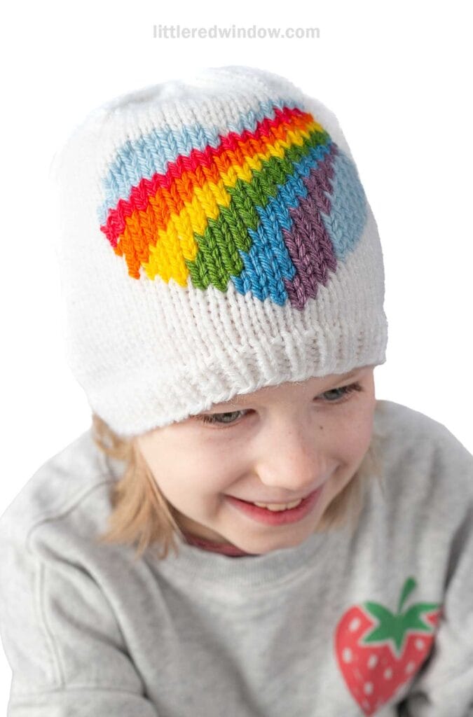 A child wearing a white knit hat with a colorful rainbow heart design. The child is smiling and wearing a light gray shirt with a strawberry graphic on it.