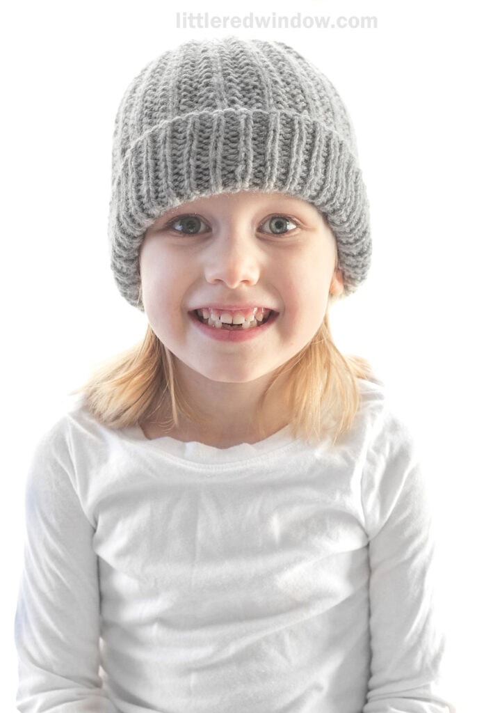 smiling girl looking at the camera and wearing a light gray ribbed knit hat with folded up brim