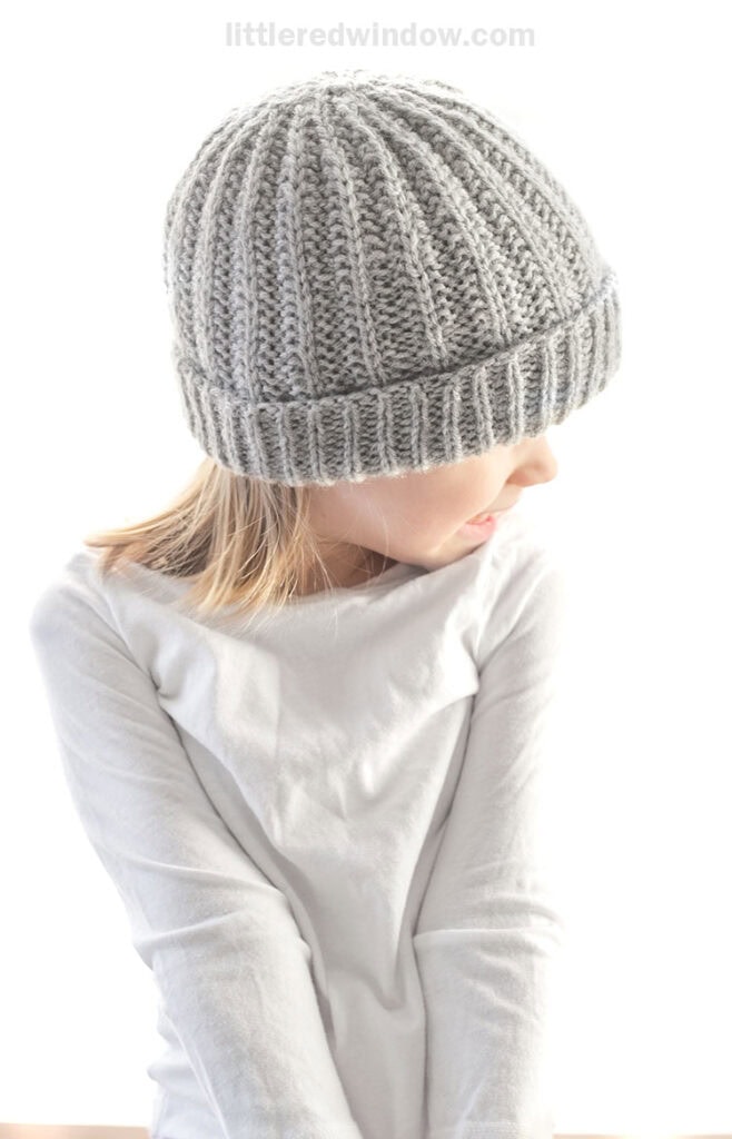 girl in white shirt wearing light gray reversible rib hat and looking off to the right with her shoulders shrugged
