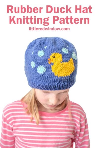 Girl wearing a blue knitted hat featuring a yellow rubber duck and bubbles pattern. She's dressed in a pink and white striped shirt. Text above reads, "Rubber Duck Hat Knitting Pattern" and the website "littleredwindow.com".