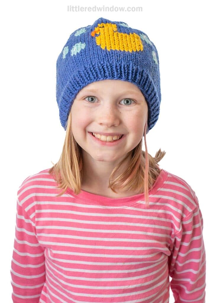 Girl smiling, wearing a blue knit beanie with a yellow duck design and light blue bubble patterns. She's dressed in a pink and white striped shirt against a white background.