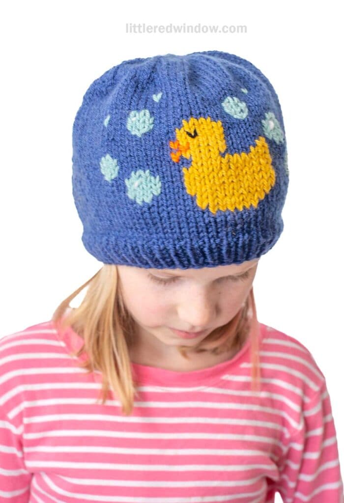 A child wearing a blue knit hat with a yellow duck and bubble design. The child is looking down, wearing a pink and white striped shirt.