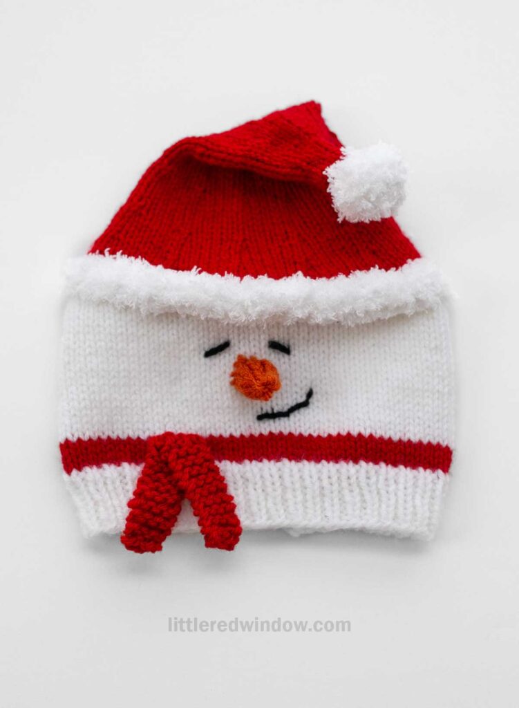 A charming Santa snowman hat, this knitted creation resembles a snowman with a festive red and white top and pom-pom. It boasts an embroidered face with eyes and a carrot nose, complemented by a red scarf detail.
