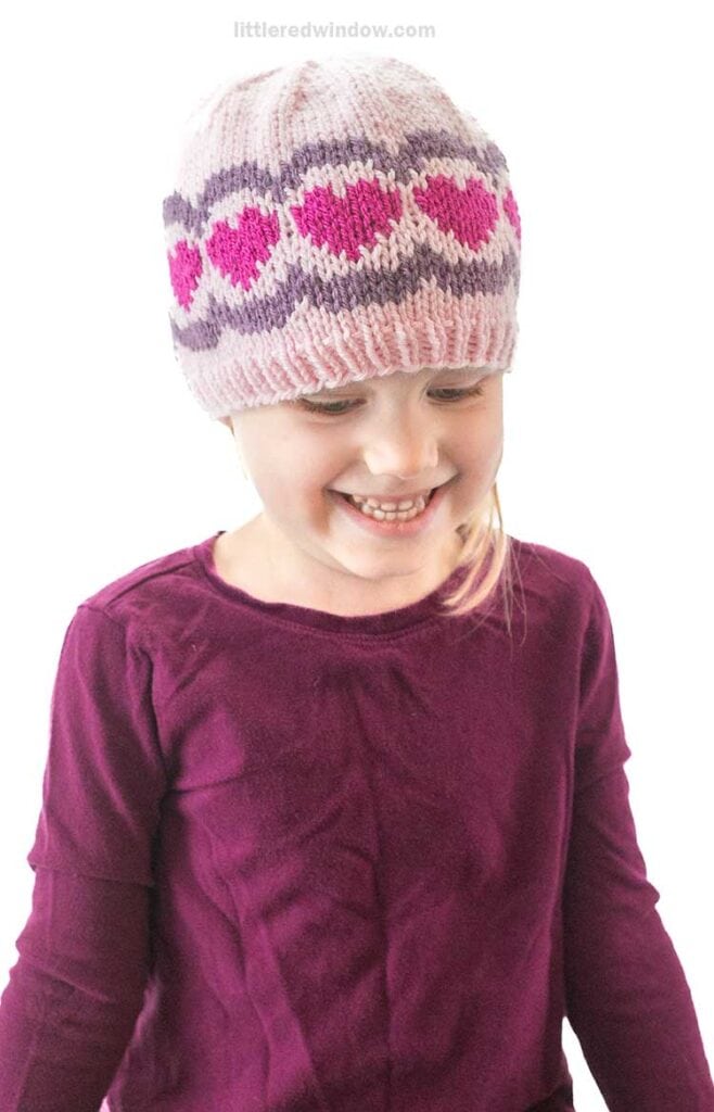 A smiling child wearing a scalloped heart hat adorned with pink and purple patterns. The child is dressed in a long-sleeved, maroon top and looks down slightly, exuding warmth and innocence.