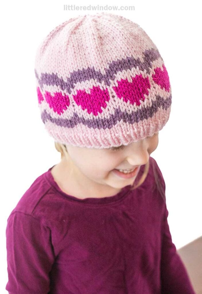 A young child wearing a scalloped heart hat adorned with purple and pink patterns smiles while looking down. They are dressed in a purple top, and the background is plain white.