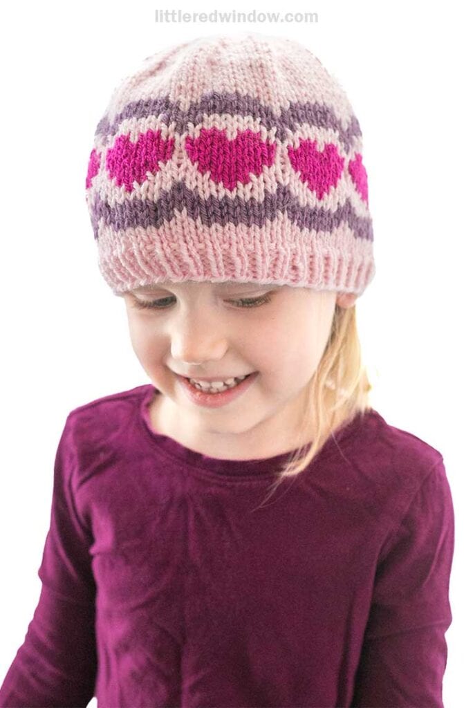 A young child smiles while looking down, wearing a scalloped heart hat with pink and purple patterns. Dressed in a matching purple top, the child stands against a plain white background.