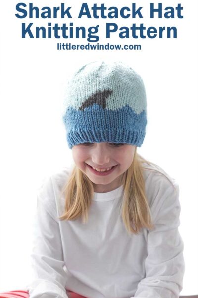 Smiling girl looking down wearing a white shirt and a knit hat with medium blue waves with a shark fin in front of a white background