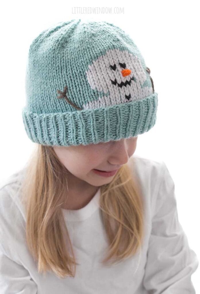 Blond girl in a white shirt wearing a light blue knit hat with a smiling snowman face peeking out over the folded brim smiling and looking down and to the right in front of a white background