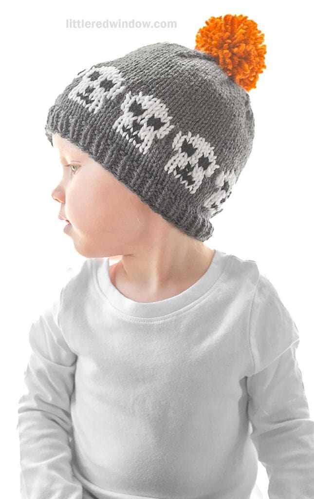 A young child is wearing a gray knitted beanie with a pattern of white skulls around the brim. The beanie has an orange pom-pom on top. The child is dressed in a white long-sleeve shirt and is looking to the side. The background is plain white.