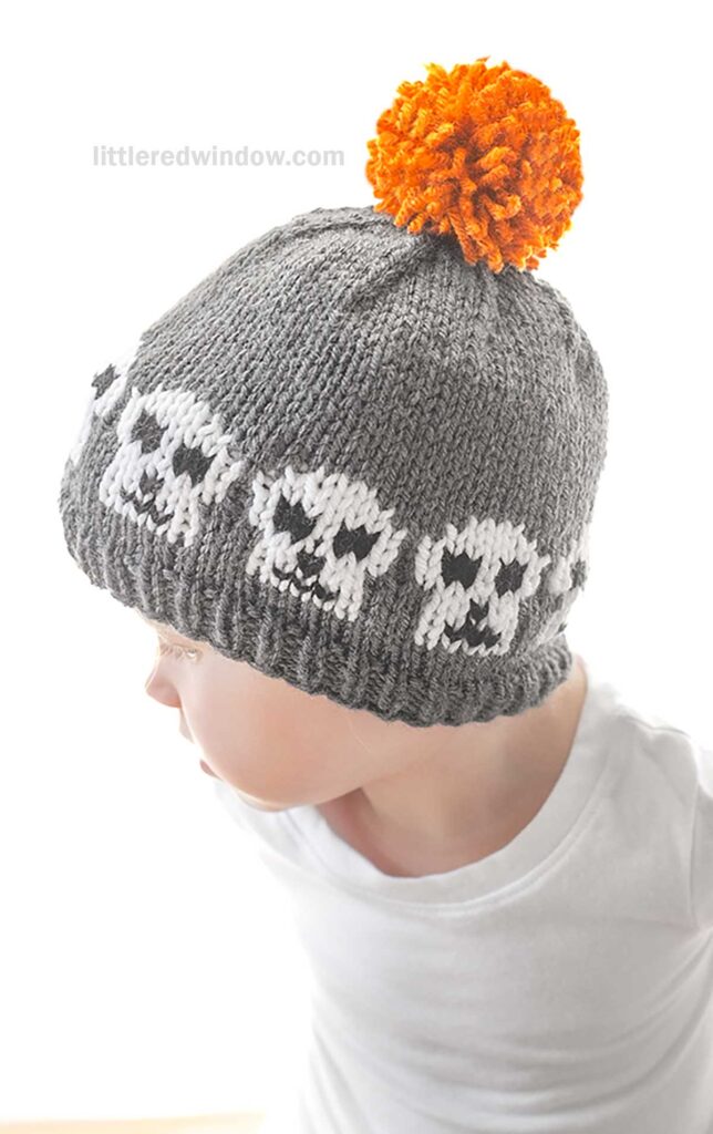 A young child wearing a gray knitted hat with a white skull pattern and an orange pom-pom on top. The child is looking down and to the side, dressed in a white shirt, against a plain white background. The watermark
