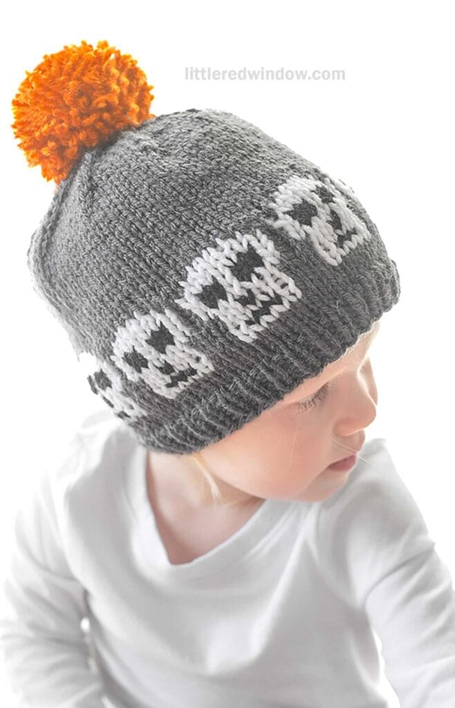 A baby wearing a gray knit hat with white skull designs around it and an orange pom-pom on top. The baby is dressed in a white shirt and is looking downward. The background is white.
