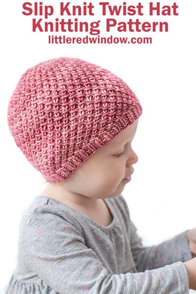 Baby in gray shirt wearing a medium pink hand dyed knit hat with a swirling slip knit twist pattern reaching off to the right in front of a white background