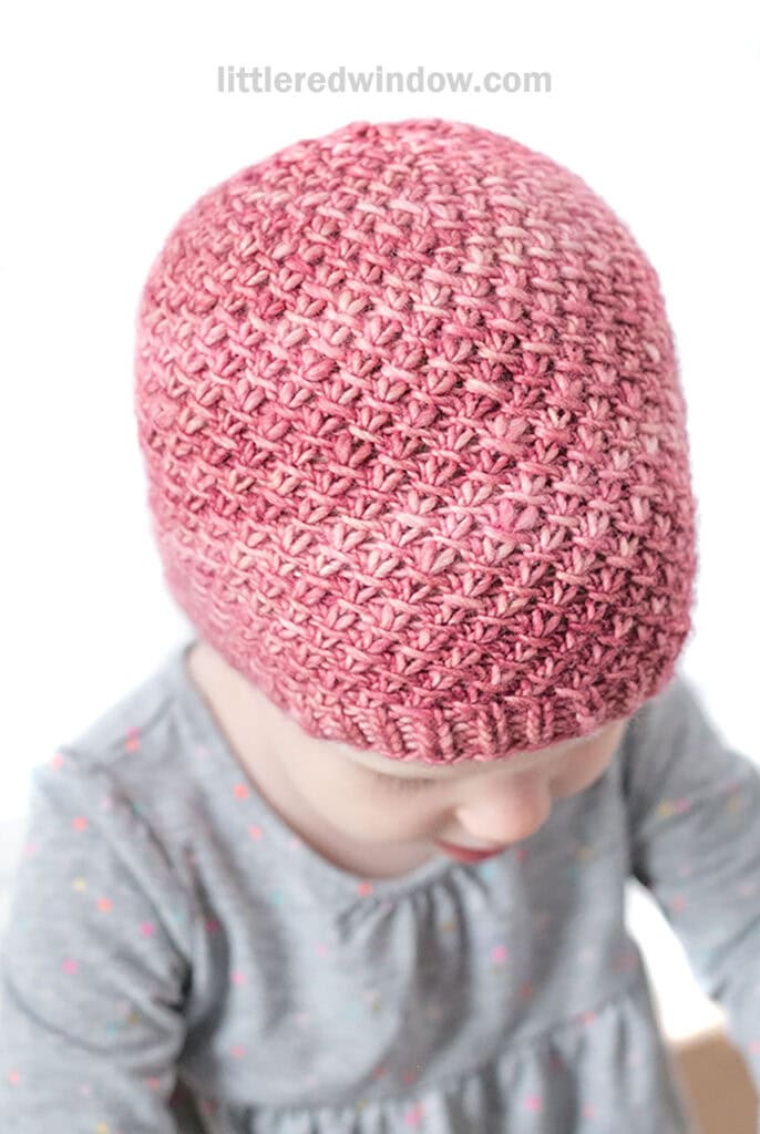Baby in gray shirt wearing a medium pink hand dyed knit hat with a swirling slip knit twist pattern looking down at their lap and to the right in front of a white background
