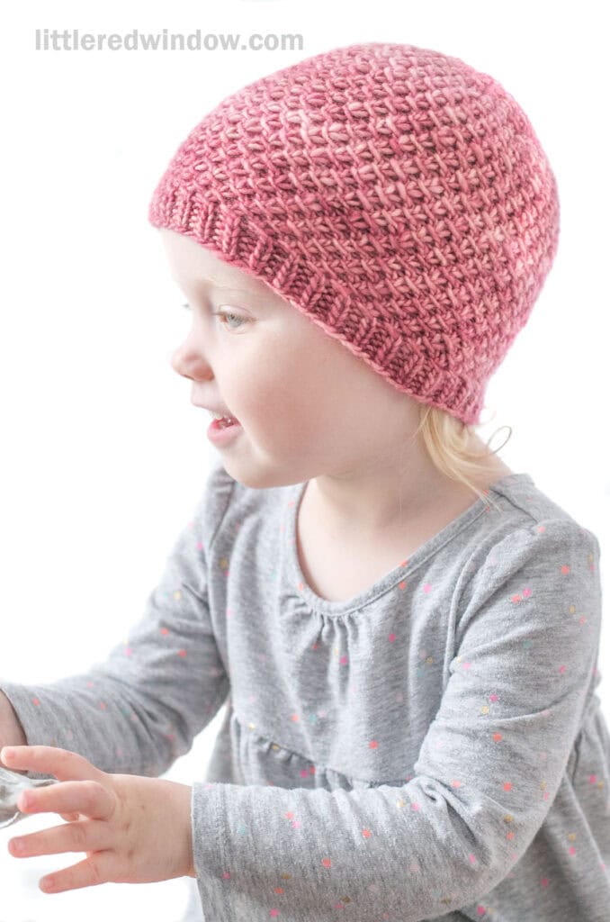 Baby in gray shirt wearing a medium pink hand dyed knit hat with a swirling slip knit twist pattern reaching off to the left in front of a white background