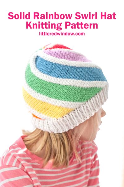 A child wearing a knitted hat with a rainbow swirl pattern and a white brim. The child is dressed in a pink and white striped shirt. Text above reads, "Solid Rainbow Swirl Hat Knitting Pattern" from "littleredwindow.com".