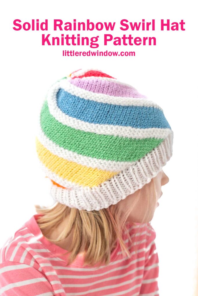 A child wearing a knitted hat with a rainbow swirl pattern and a white brim. The child is dressed in a pink and white striped shirt. Text above reads,