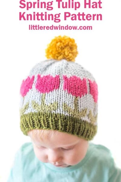 A baby wearing a knitted hat with pink tulip patterns, green leaves, and a yellow pom-pom on top. The text above reads, "Spring Tulip Hat Knitting Pattern littleredwindow.com.