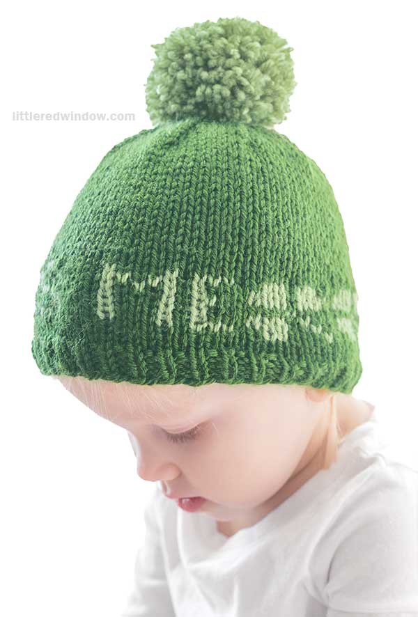A young child, sporting a green St. Patrick's Day hat with a pom-pom, peers downward. The hat features festive white letters knitted around its edge, perfectly matching the child's crisp white shirt.
