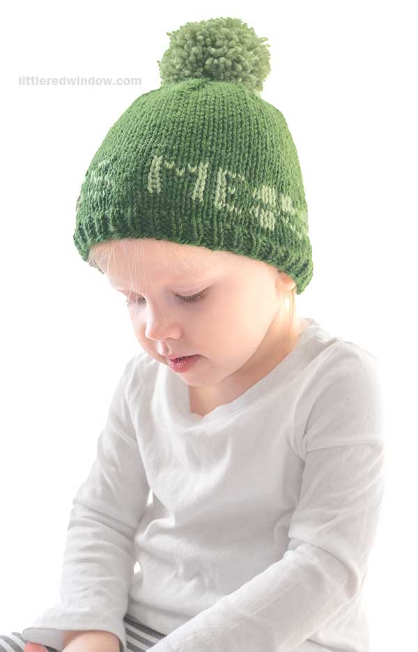 A young child with short blonde hair is wearing a green St. Patrick's Day hat adorned with a pom-pom and a white, long-sleeve shirt. The plain, well-lit background adds to the charm as the child looks downward, lost in thought.