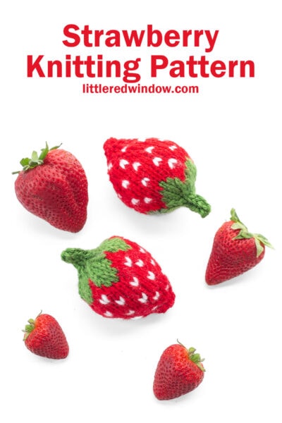 two red knit strawberries with green leaves on a white tabletop with three real strawberries viewed from above