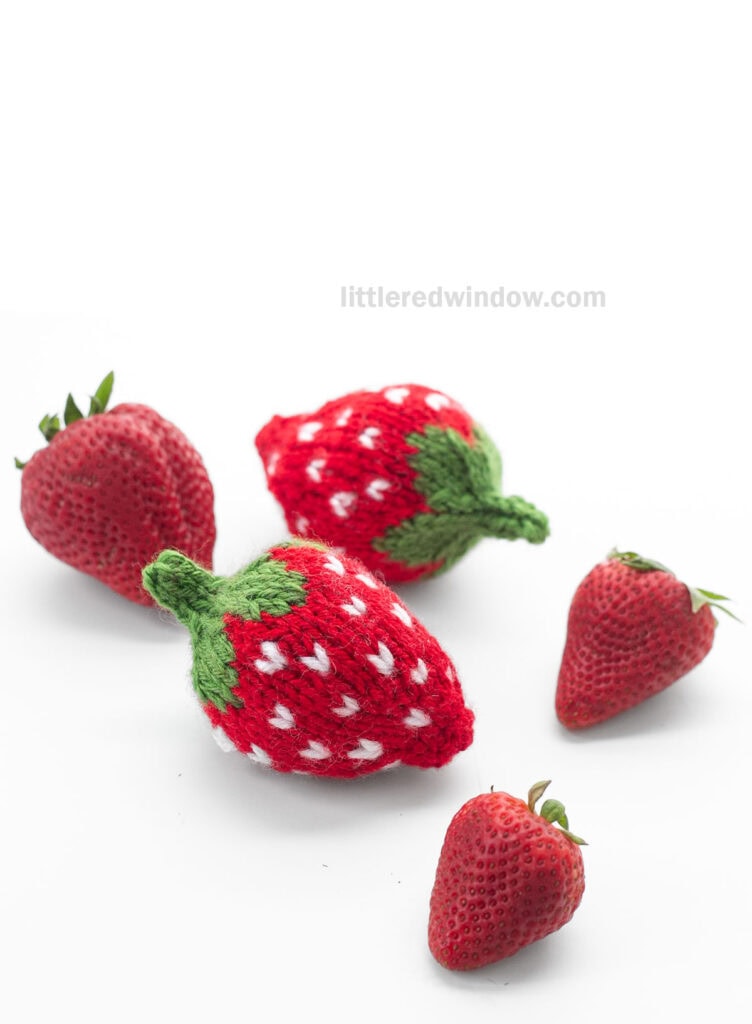two red knit strawberries with green leaves on a white tabletop with three real strawberries viewed from down low