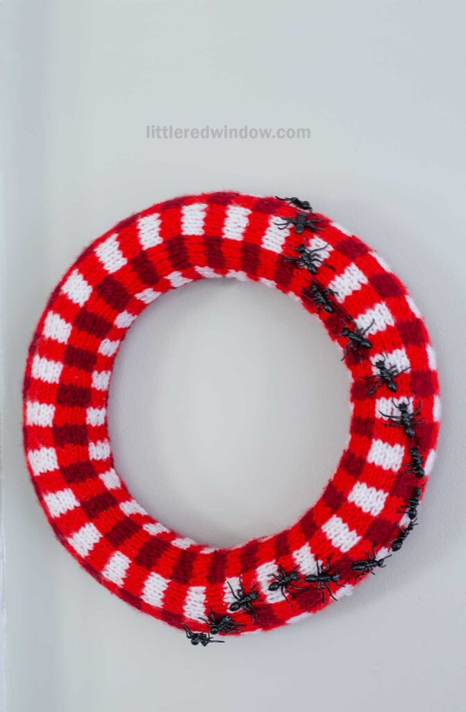 A red and white plaid knitted wreath hangs on a wall, with several plastic black ants attached along one side.