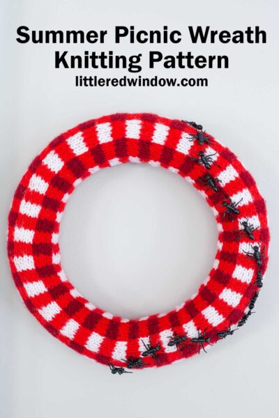 A red and white plaid knitted wreath resembling a picnic blanket, decorated with several plastic ants, displayed on a white background. Text above reads: "Summer Picnic Wreath Knitting Pattern littleredwindow.com".