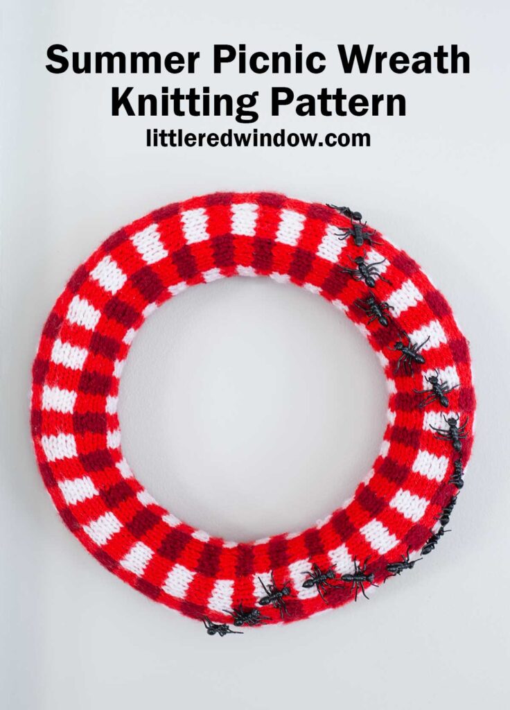 A red and white plaid knitted wreath resembling a picnic blanket, decorated with several plastic ants, displayed on a white background. Text above reads: 