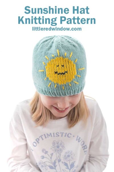 smiling child in white shirt with blonde hair wearing a sky blue knit hat with a bright yellow smiling sunshine on the front looking down at their lap
