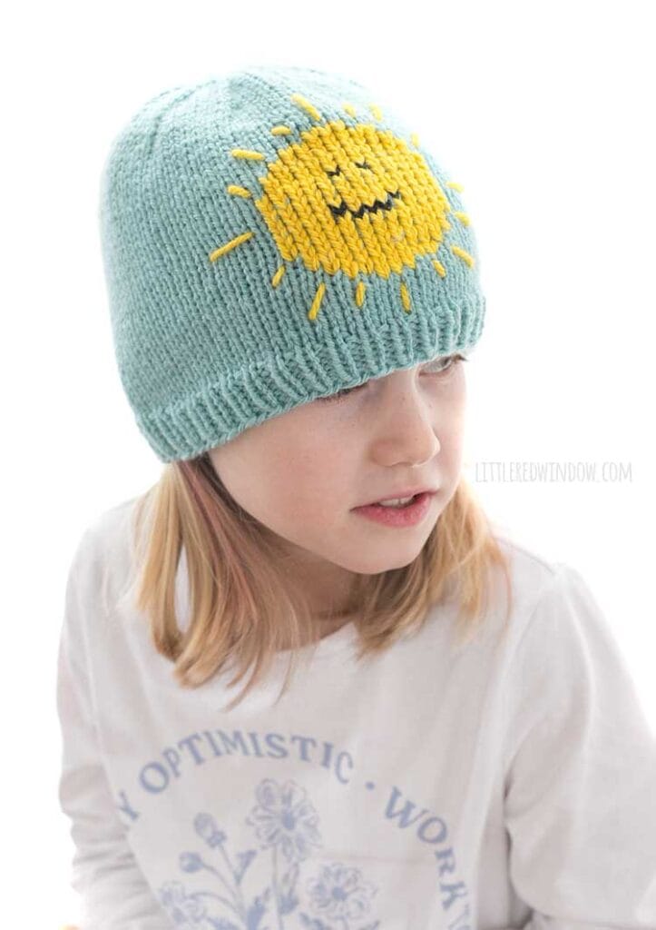 child in light sky blue knit hat with yellow smiling sunshine on the front looking over their left shoulder