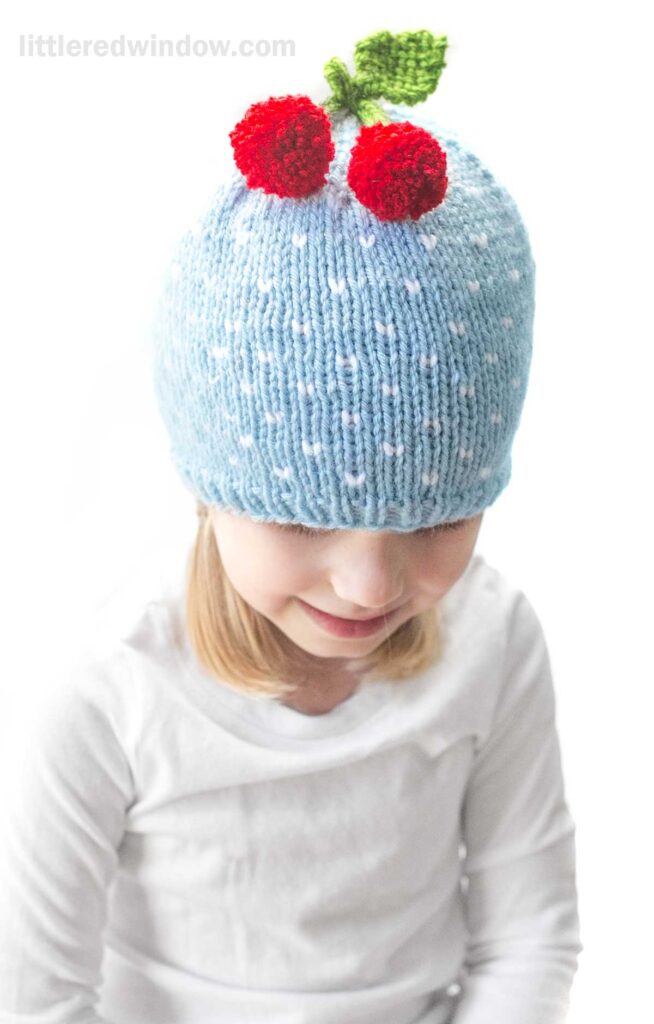 A young child wearing a cherry hat, specifically a light blue knit hat adorned with two red cherries on green stems. The child is dressed in a white shirt and is looking downward. The background is plain white.
