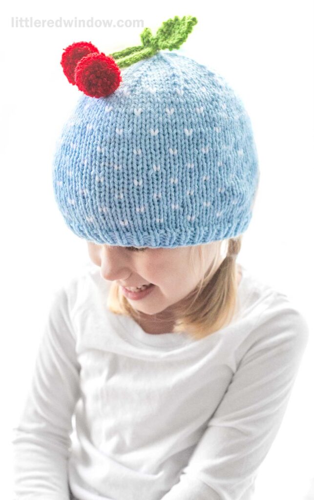 A young girl wearing a white shirt and a cherry hat adorned with red cherries on top. She is smiling and looking down. The background is bright and mostly white.