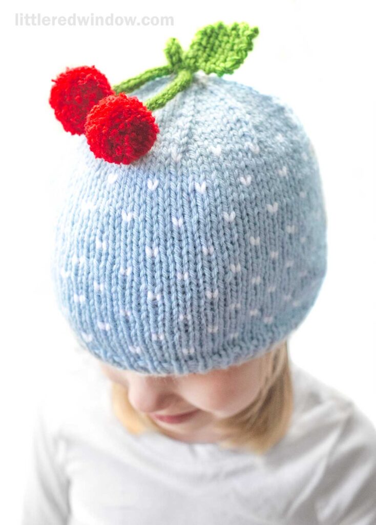 A child wears a charming cherry hat: a light blue knitted beanie with white polka dots. Two red pom-pom cherries with green leaves adorn the top. The child's face is partially visible, looking down, against a white background.