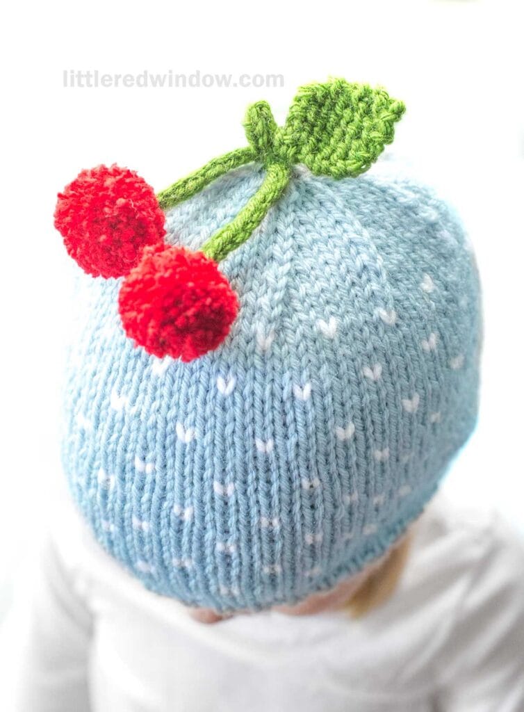 A child wearing a light blue cherry hat decorated with tiny white polka dots. The hat is adorned with two bright red cherries and green leaves knitted on top. The background is white, and the text