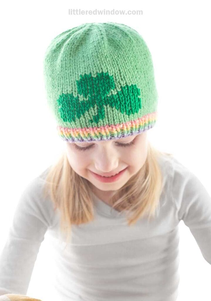 A young girl is wearing a green knit hat featuring a charming shamrock knitting pattern. She is smiling and dressed in a white long-sleeve shirt. The plain white background emphasizes her as the focal point of the image.