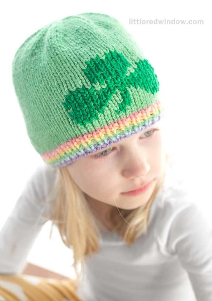 A young girl dons a light green knitted hat featuring a charming shamrock design. The hat, adorned with a rainbow band at the edge, showcases a delightful knitting pattern. Sporting a white long-sleeved shirt, she gazes slightly to the side.